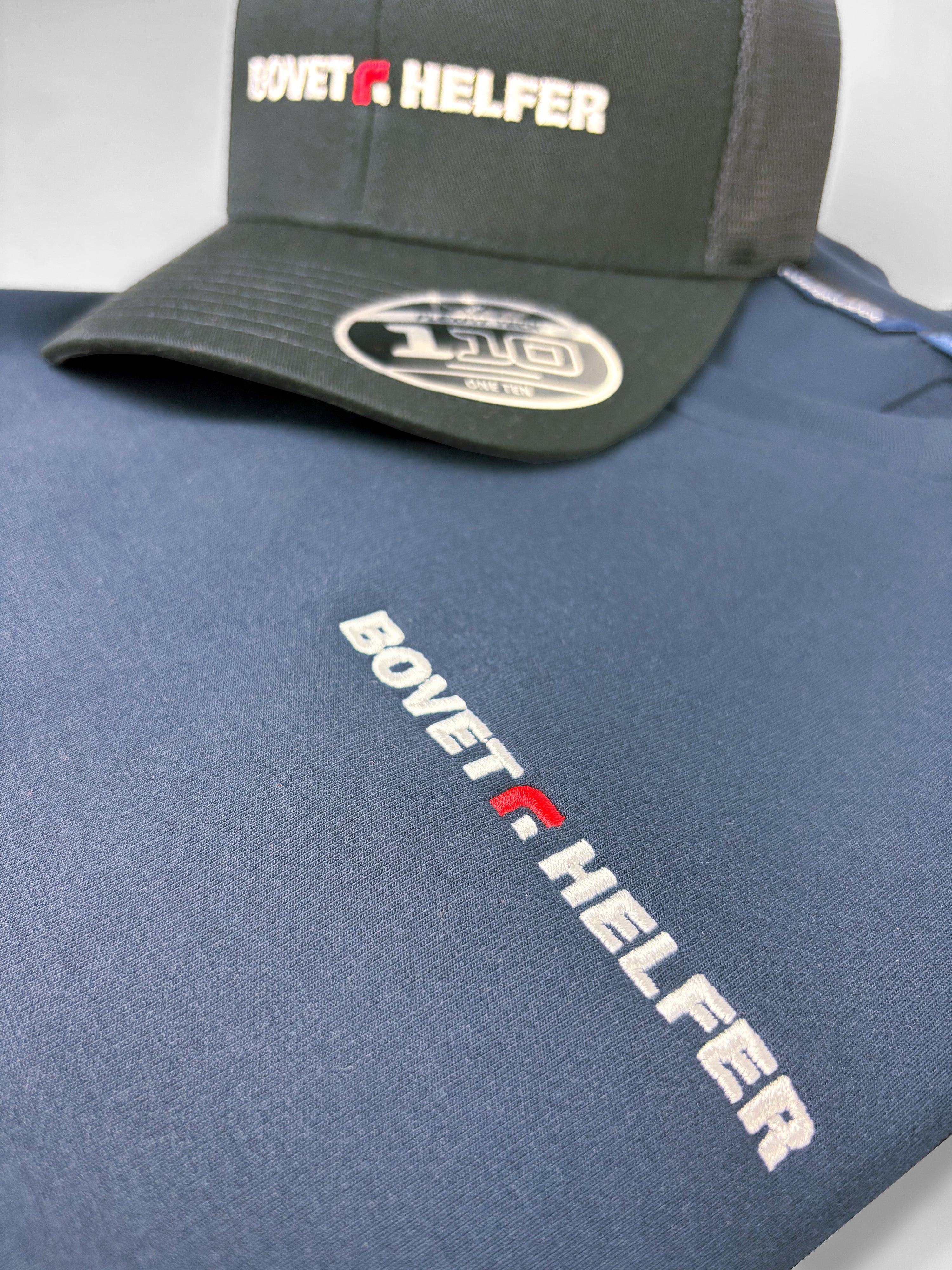 Broderie verticale Bovet+Helfer en blanc et rouge sur un sweat bleu marine, casquette noire à proximité.